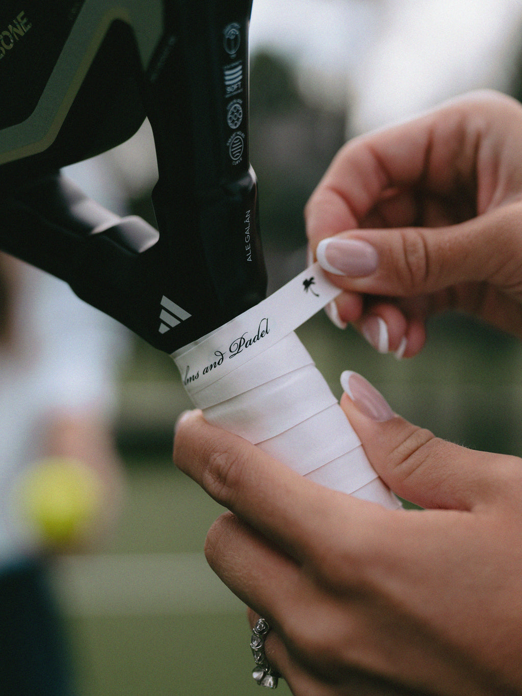Close-up of a padel player wrapping a white overgrip tape with black script text on a black padel racket handle, with hands and a blurred padel court in the background.