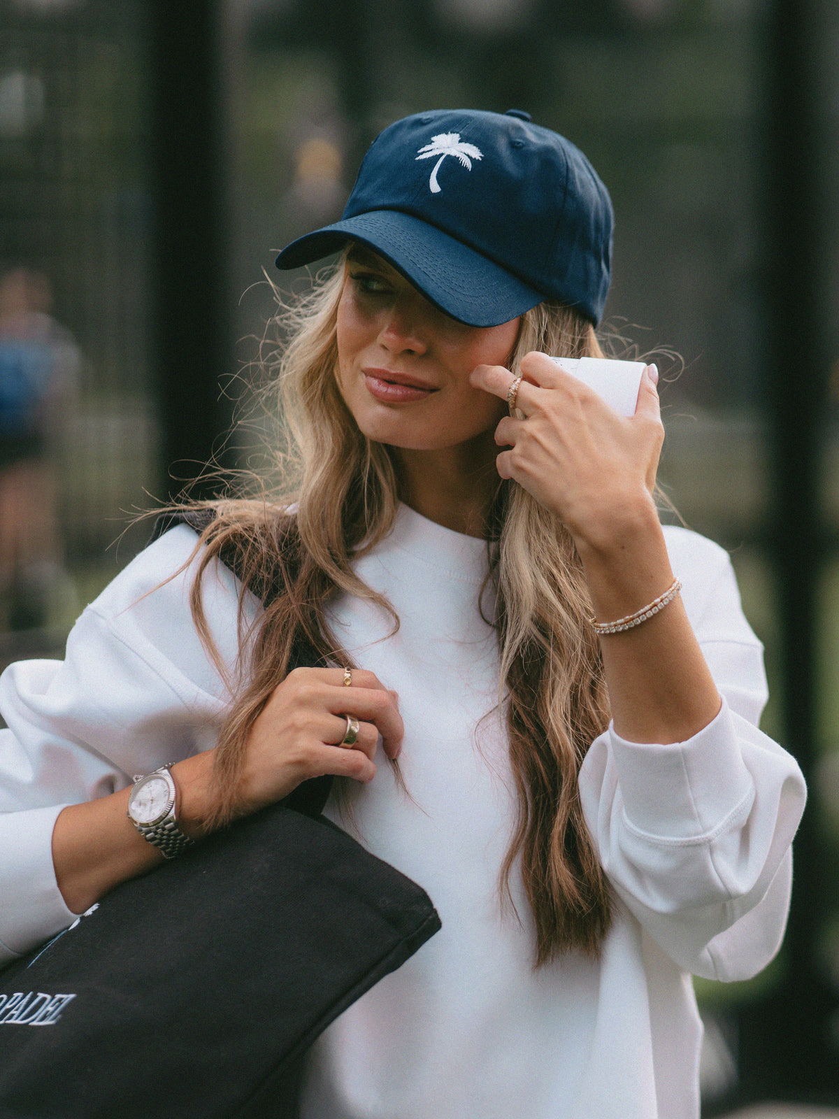 A woman wearing a navy blue baseball cap with a white palm tree logo, a white sweatshirt, and carrying a black tote bag, photographed in an outdoor street setting.