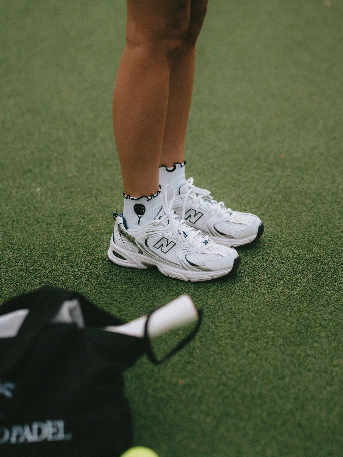 White New Balance 990v5 sneakers worn with white padel racket socks on a green turf court, with a black Padel bag in the foreground.