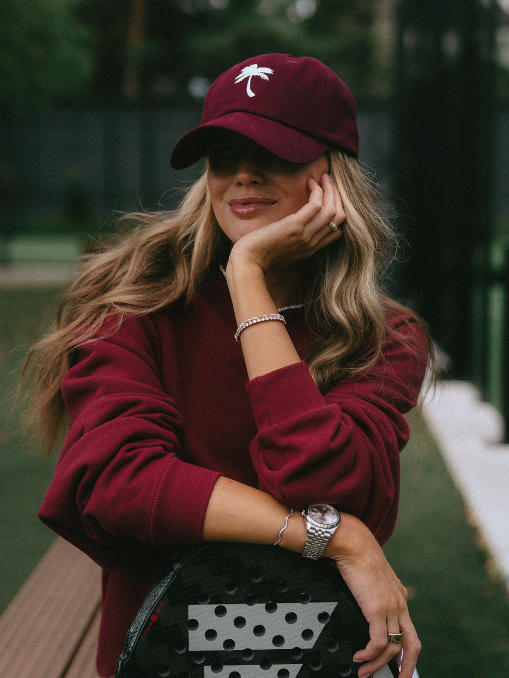 A woman wearing a burgundy long-sleeve sweatshirt and a matching burgundy baseball cap with a white palm tree logo, leaning on a padlel racket in an outdoor park setting.