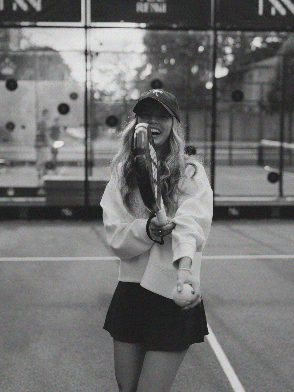 A woman on a tennis court wearing a white sweatshirt, black tennis skirt, and a dark baseball cap, holding a padel racket and ball in a black and white photograph.