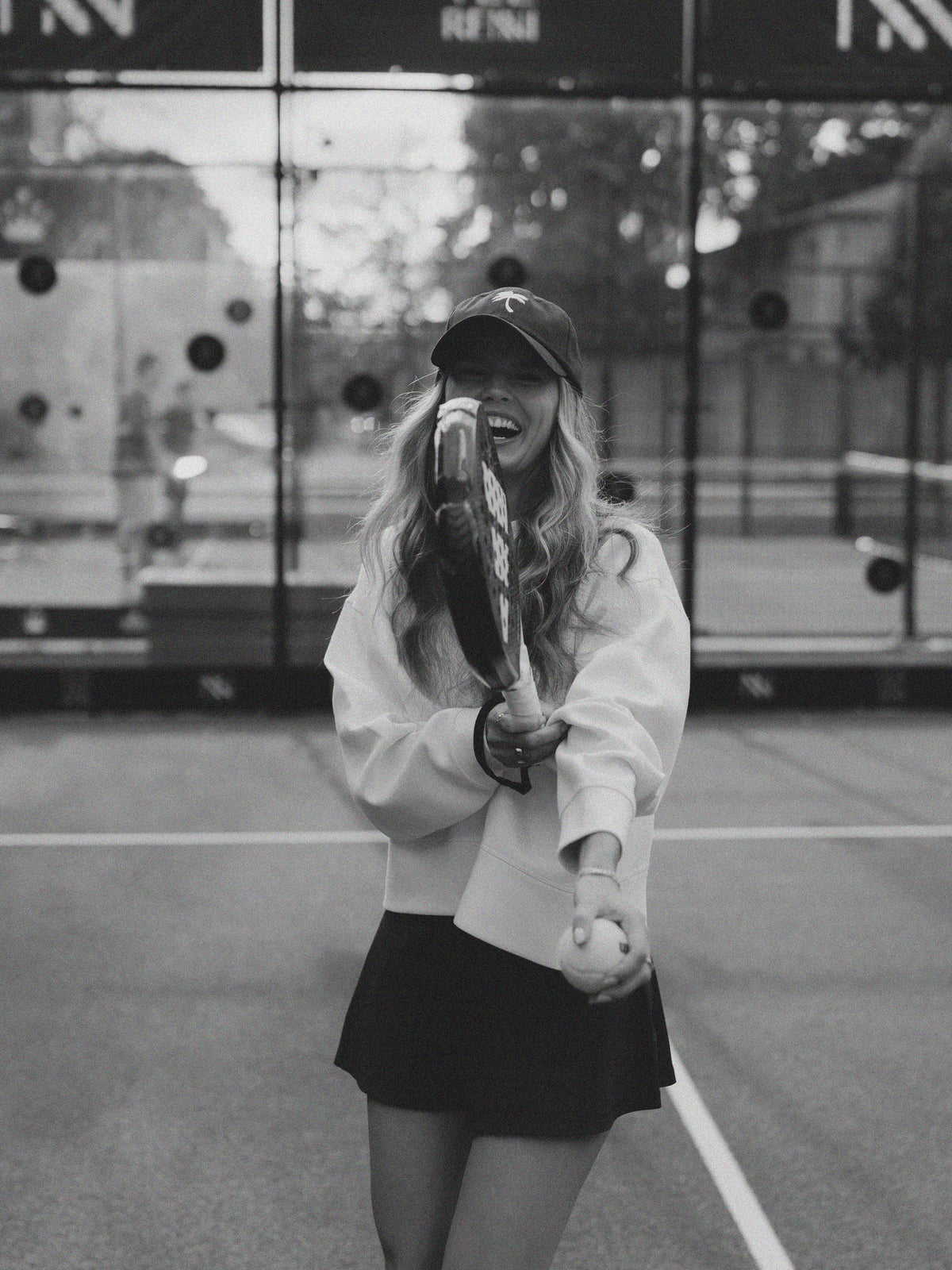 A woman on a tennis court wearing a white sweatshirt, black tennis skirt, and a dark baseball cap, holding a padel racket and ball in a black and white photograph.
