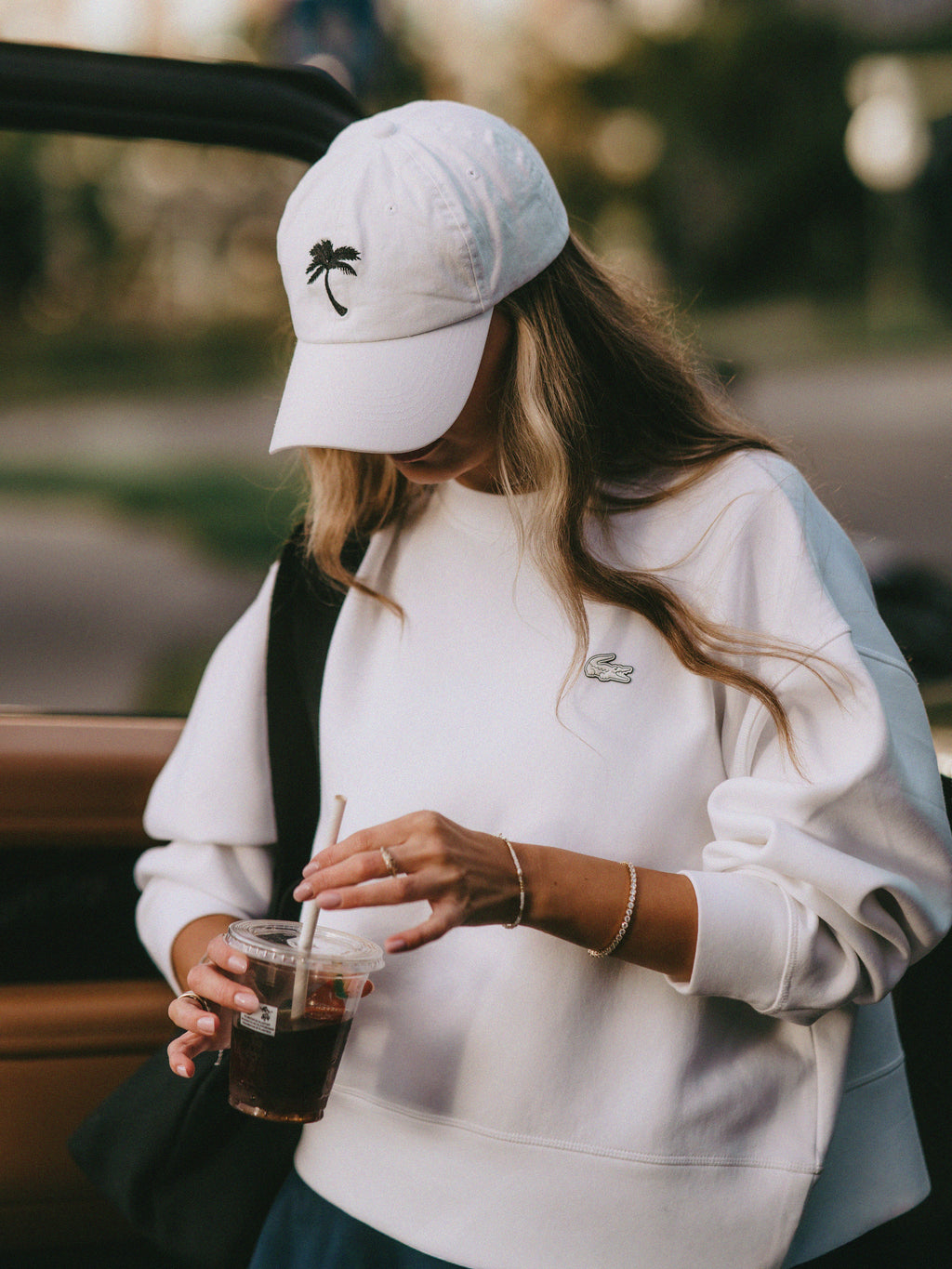 A woman wearing a white Lacoste sweatshirt and a white baseball cap with a black palm tree logo, holding an iced coffee in a sunny outdoor setting.