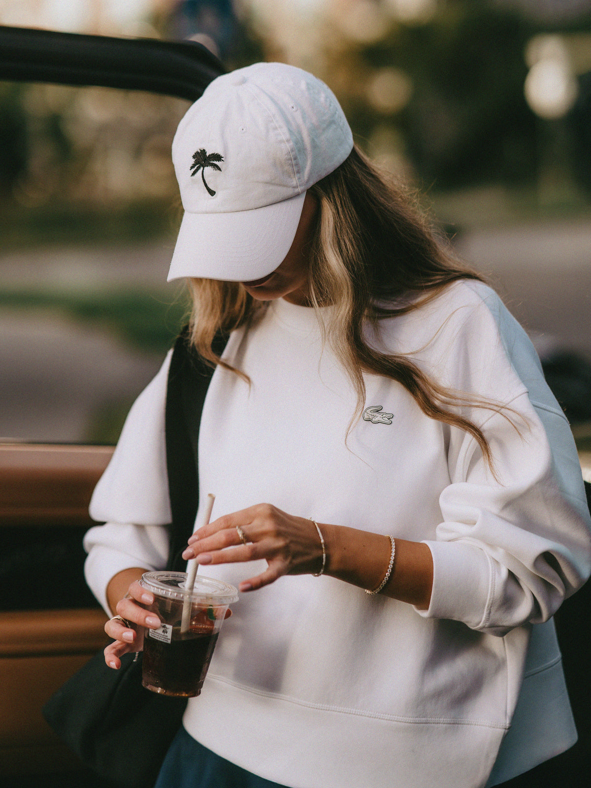 A woman wearing a white Lacoste sweatshirt and a white baseball cap with a black palm tree logo, holding an iced coffee in a sunny outdoor setting.